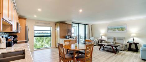A spacious living area with a dining table, chairs, and a sofa. There is a kitchen area with cabinets and a countertop. A large window provides a view of greenery outside.