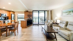A spacious living area with a dining table and chairs, a sofa, and a television. There is a kitchen area with wooden cabinets and a refrigerator. A balcony with a view of the water is visible. - Marco Island Waterfront Condo w/ Community Pool! (Marco Island)