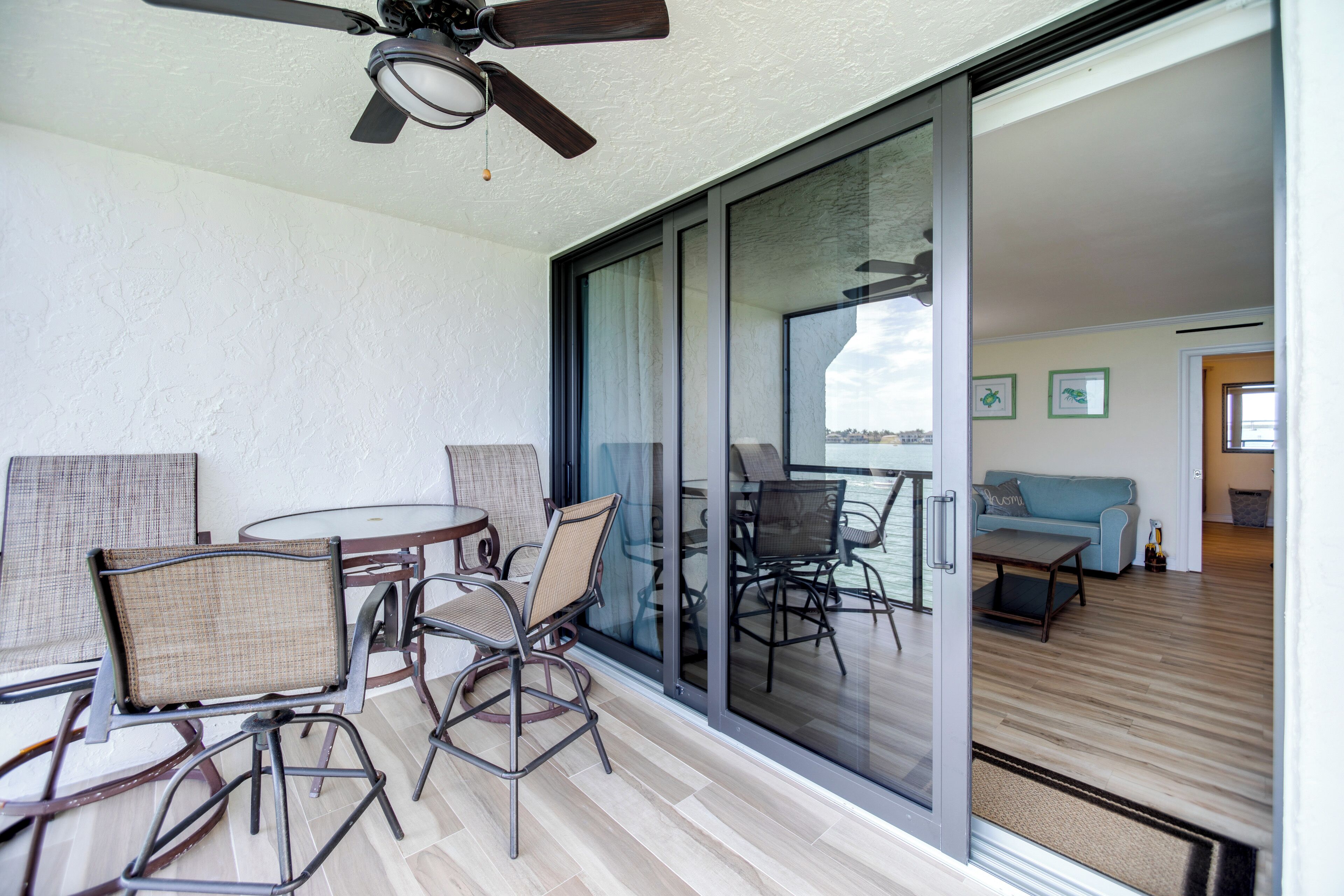 Ein Balkon mit Deckenventilator, eine Glastür zu einem Wohnbereich mit Sofa, Couchtisch und Fernseher.