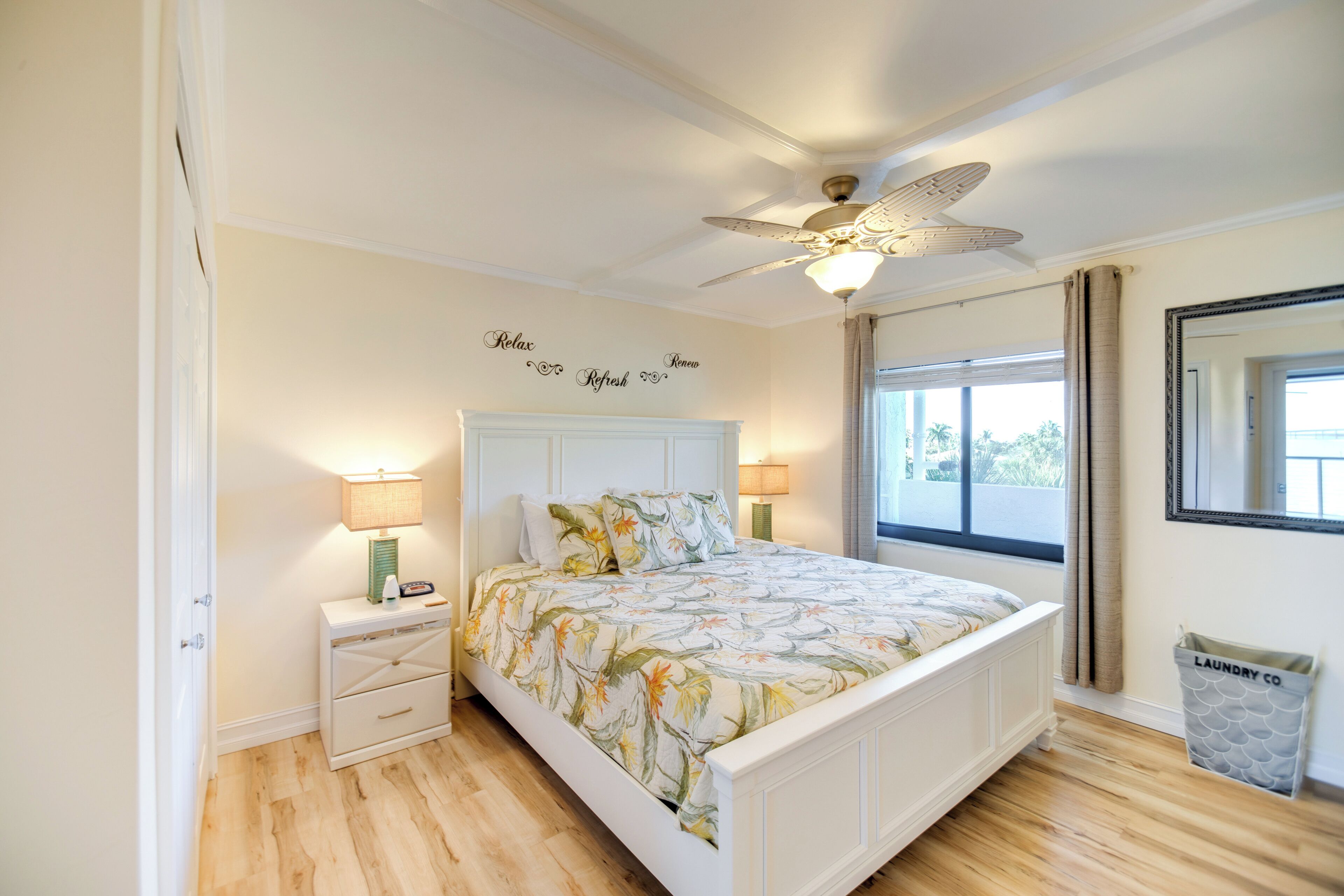 A bedroom with a bed, nightstands, a ceiling fan, a window with curtains, and a wall-mounted mirror.