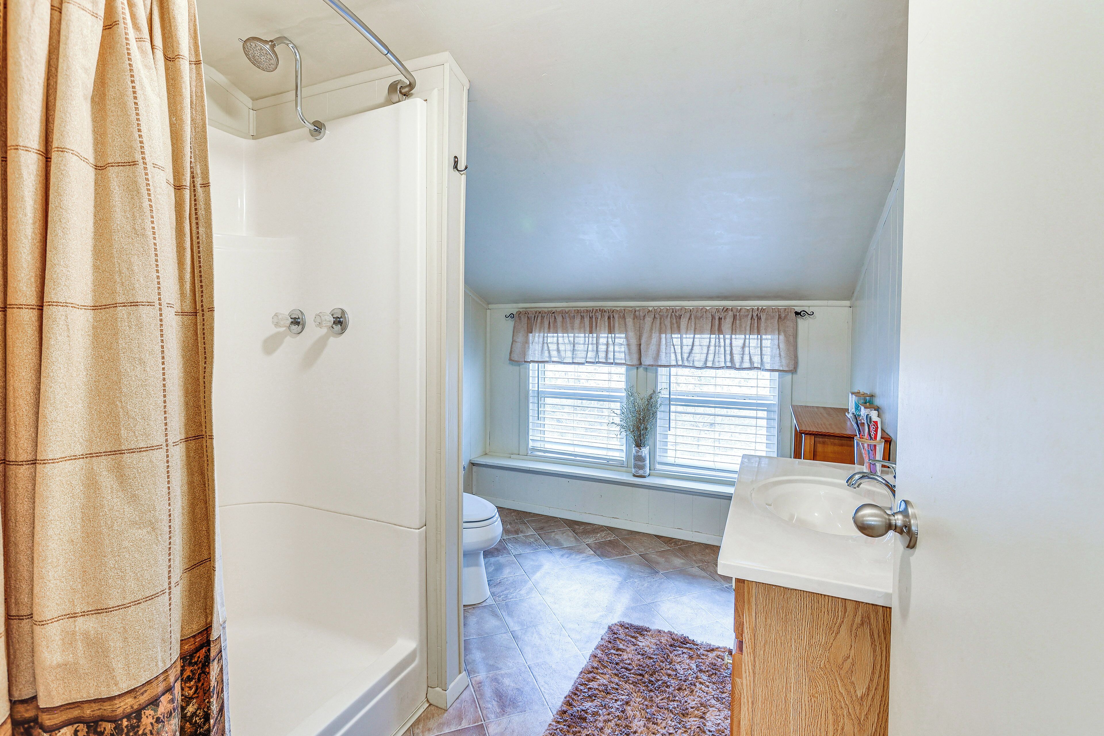 A bathroom with a shower, toilet, sink, and window with curtains.