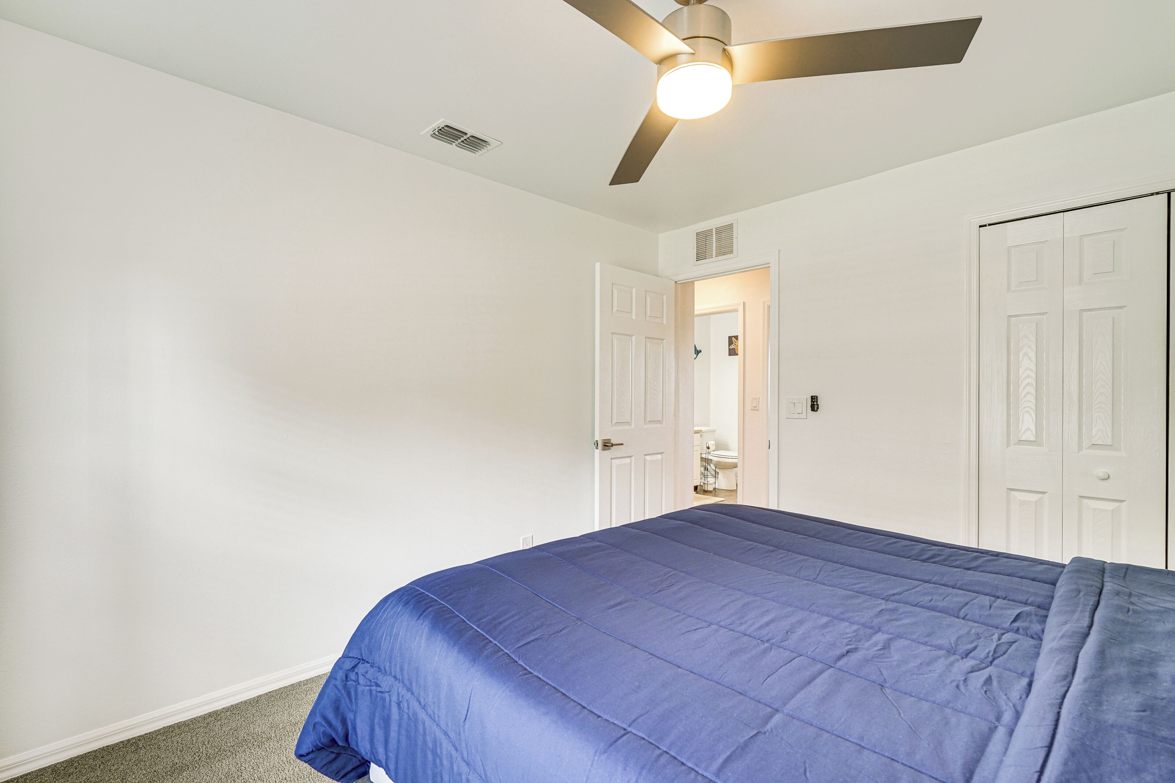 Une chambre à coucher avec un lit, des murs blancs, un ventilateur de plafond et une porte donnant sur une salle de bain.