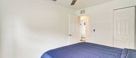 Une chambre à coucher avec un lit, des murs blancs, un ventilateur de plafond et une porte donnant sur une salle de bain.