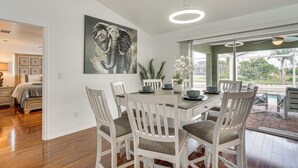 Un comedor con una mesa y sillas blancas, un cuadro grande de un elefante en la pared y vistas al exterior a través de amplias ventanas.