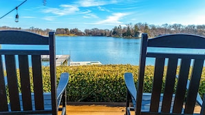 Dos sillas Adirondack mirando un lago con un muelle y un seto al frente.