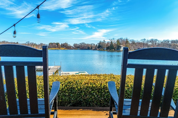 Two Adirondack chairs facing a lake with a dock and a hedge in the foreground.
