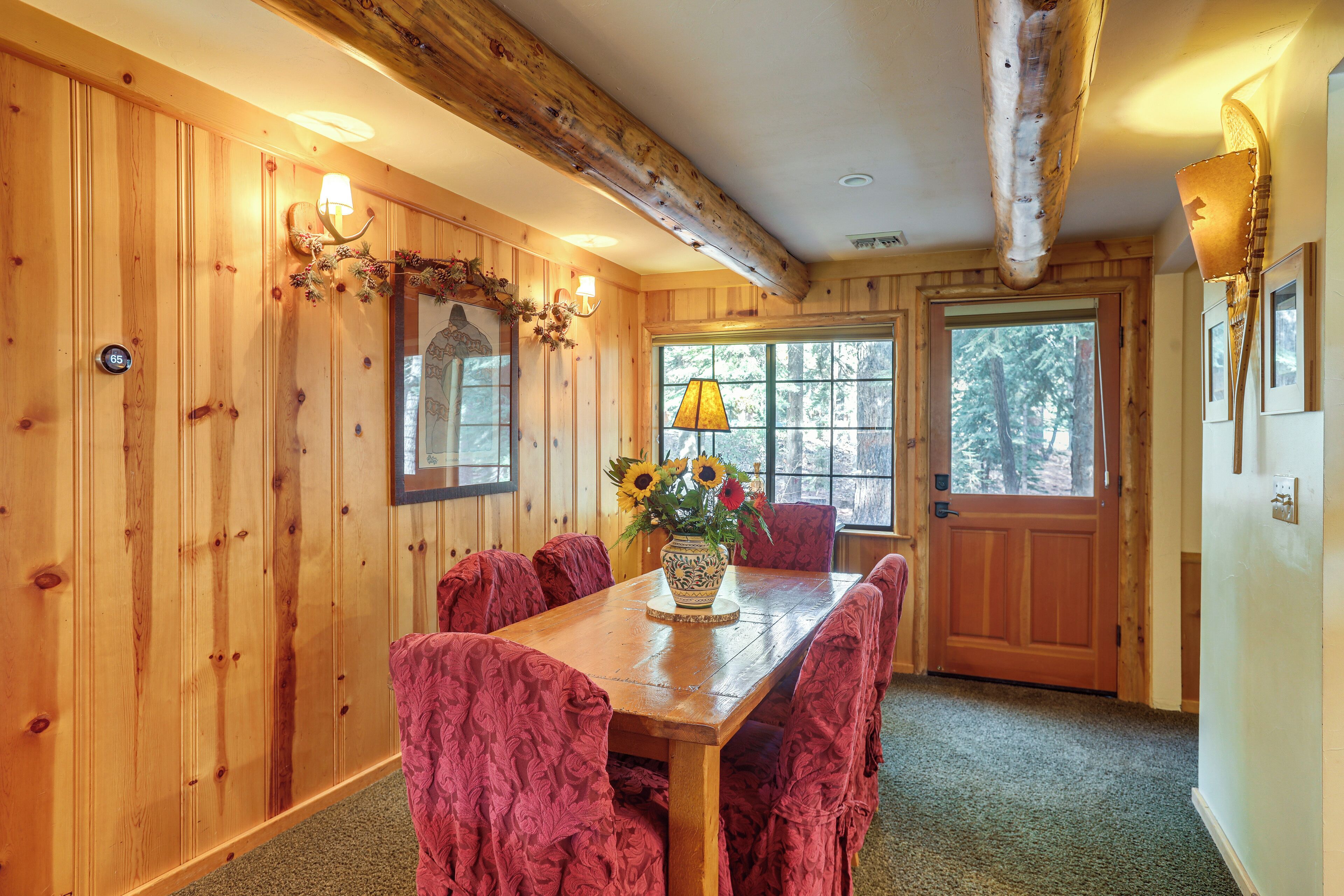 A wooden dining room with a table and chairs, a vase with flowers, and a window with a view of trees.