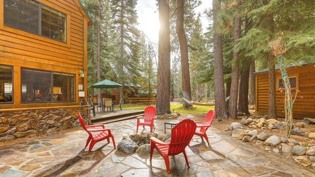 Une terrasse en pierre avec des chaises rouges et une table, entourée d’arbres et d’un chalet en bois.
