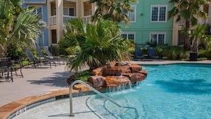 A pool with a rock waterfall feature, surrounded by lounge chairs and palm trees. - Gulf & Pool view, Washer & Dryer, Families Love it (Galveston)