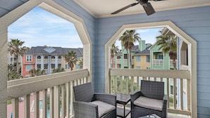 A balcony with wicker furniture and a ceiling fan, overlooking colorful buildings and palm trees. - Gulf & Pool view, Washer & Dryer, Families Love it (Galveston)