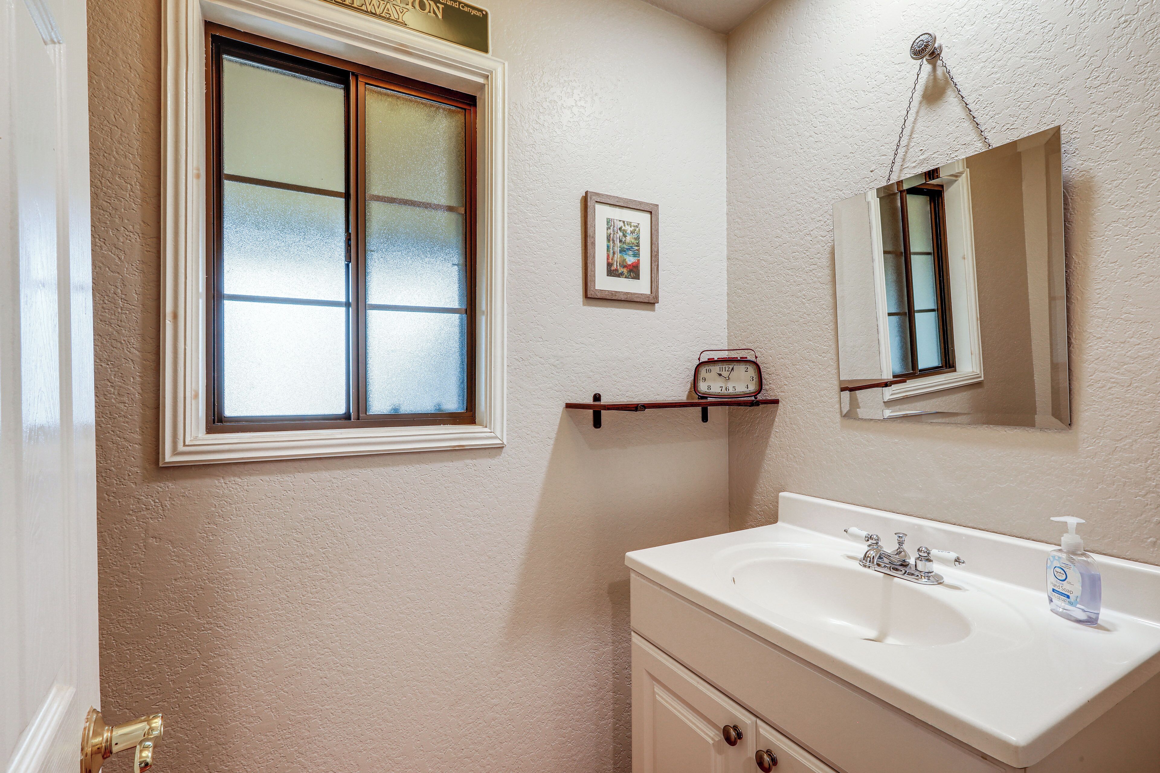 A bathroom with a white sink, a mirror, a clock, and a window with frosted glass.