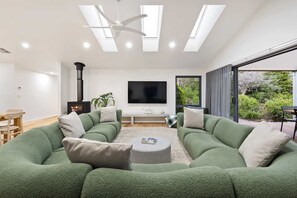 A modern living room with a green sectional sofa, a flat-screen TV, a fireplace, and large windows offering a view of greenery.