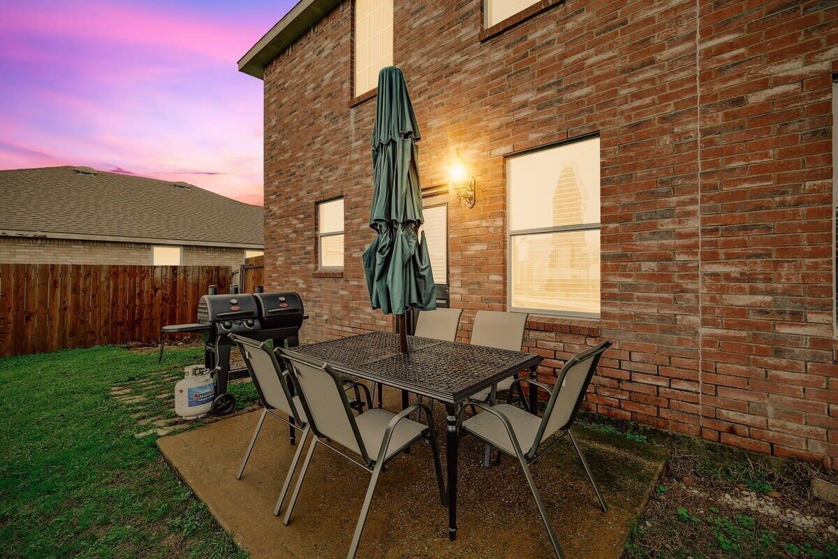 A backyard patio with a brick house, a grill, a table with chairs, and a covered umbrella.