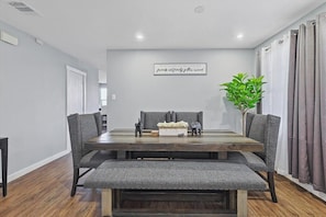 A dining area with a wooden table, grey upholstered chairs, a bench, a potted plant, and a wall-mounted sign. - 4 Bedrooms, Brick Retreat with Gameroom & Backyard (Grand Prairie)