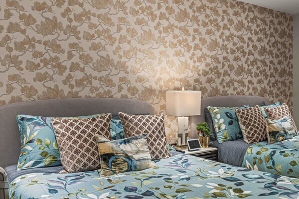 A bedroom with a floral wallpaper, a bed with patterned pillows, a bedside table with a lamp, and a laptop.
