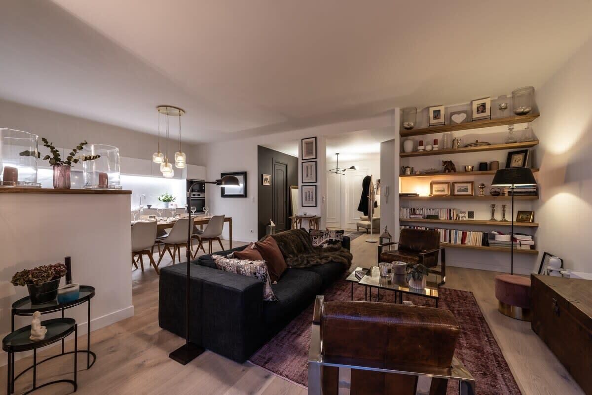 Un salon moderne avec un canapé noir, un fauteuil marron et une table basse en verre. On aperçoit une bibliothèque remplie de divers objets et, au fond, un coin repas.