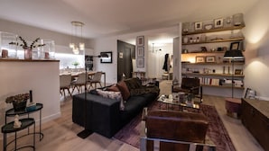 A modern living room with a black sofa, a brown armchair, and a glass coffee table. There is a bookshelf with various items and a dining area in the background. - Luxurious apartment in the heart of Annecy (Annecy)