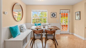 A dining area with a white bench, two wooden chairs, a table set with plates and glasses, and a large window with a view of greenery.