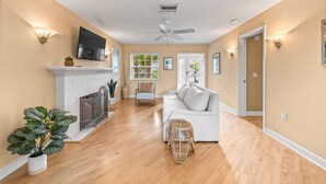 A spacious living room with a white fireplace, a ceiling fan, and a large window.