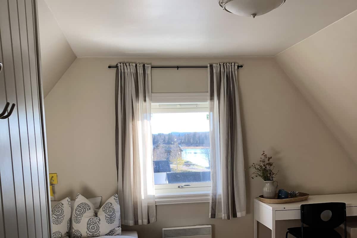 A bedroom with a bed, a desk, and a chair. There is a window with curtains, a radiator, and a view of a landscape.