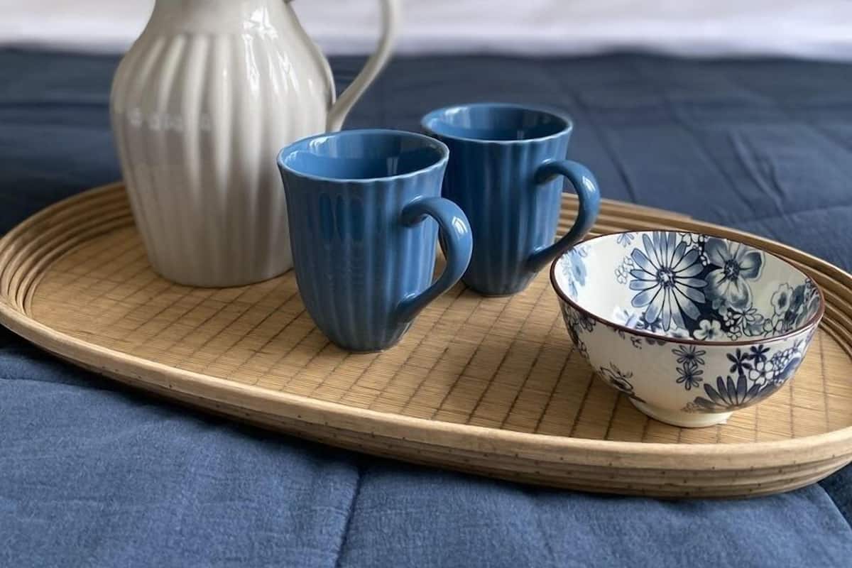 A bed with a blue bedspread, a white vase with flowers, two blue cups, and a blue and white bowl on a tray.