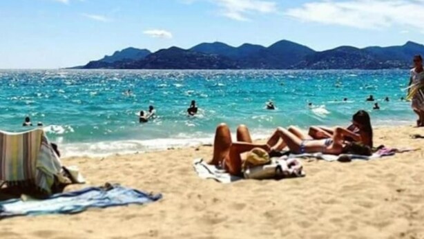 Una spiaggia con persone che prendono il sole e nuotano, con montagne sullo sfondo.