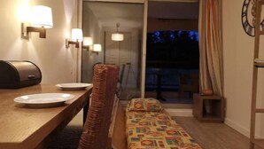 Una camera d'albergo con un tavolo da pranzo, una sedia in vimini, un letto con biancheria a motivi decorativi, una sveglia a parete e una porta scorrevole in vetro che dà su uno spazio esterno.