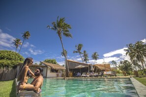 A swimming pool with lounge chairs and umbrellas, surrounded by palm trees and a thatched-roof building. - Monan Boutique Hotel  (Porto de Pedras)