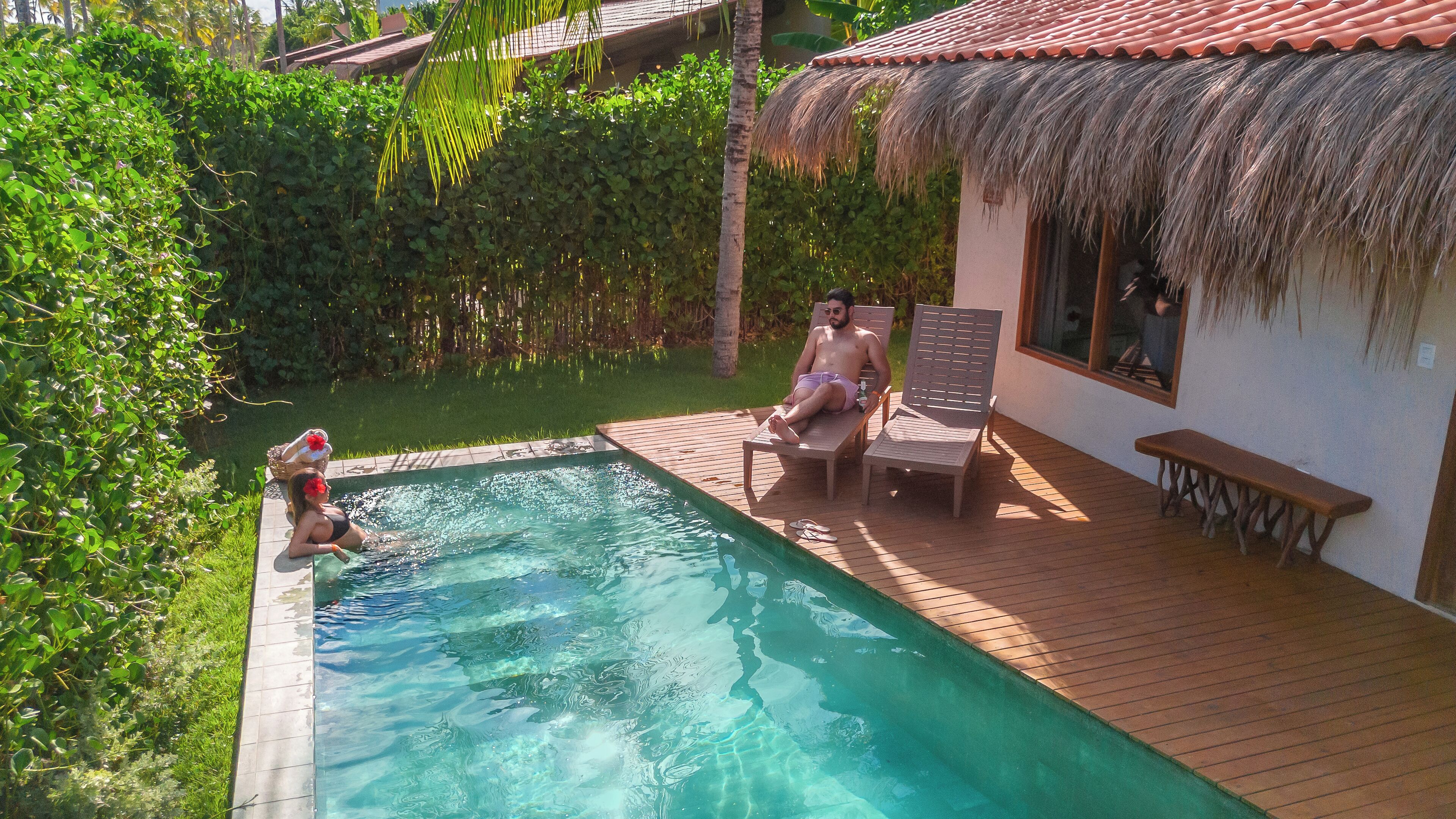 Un área de piscina con una terraza de madera, una estructura con techo de paja y abundante vegetación.