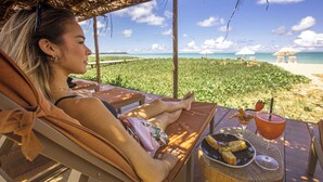 A person relaxing on a sun lounger with a beach view, a table with a drink and snacks, and a thatched roof structure. - Monan Boutique Hotel  (Porto de Pedras)