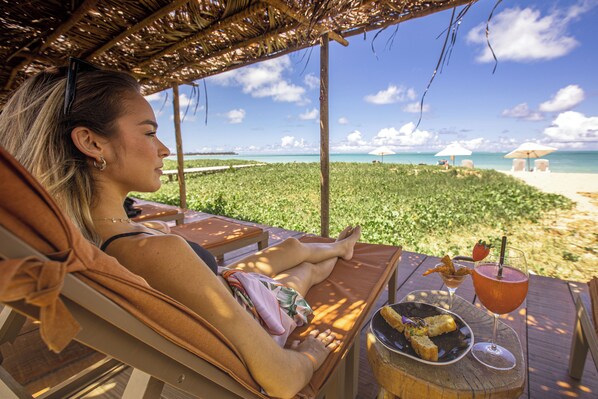 A person relaxing on a sun lounger with a beach view, a table with a drink and snacks, and a thatched roof structure. - Monan Boutique Hotel  (Porto de Pedras)