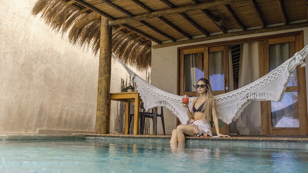 A woman sitting in a hammock by a pool with a thatched roof and wooden doors. - Monan Boutique Hotel  (Porto de Pedras)