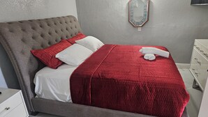 Ein ordentlich bezogenes Bett mit roter Decke und Kissen, ein grau gepolsterter Kopfteil und ein Nachttisch mit Schubladen.