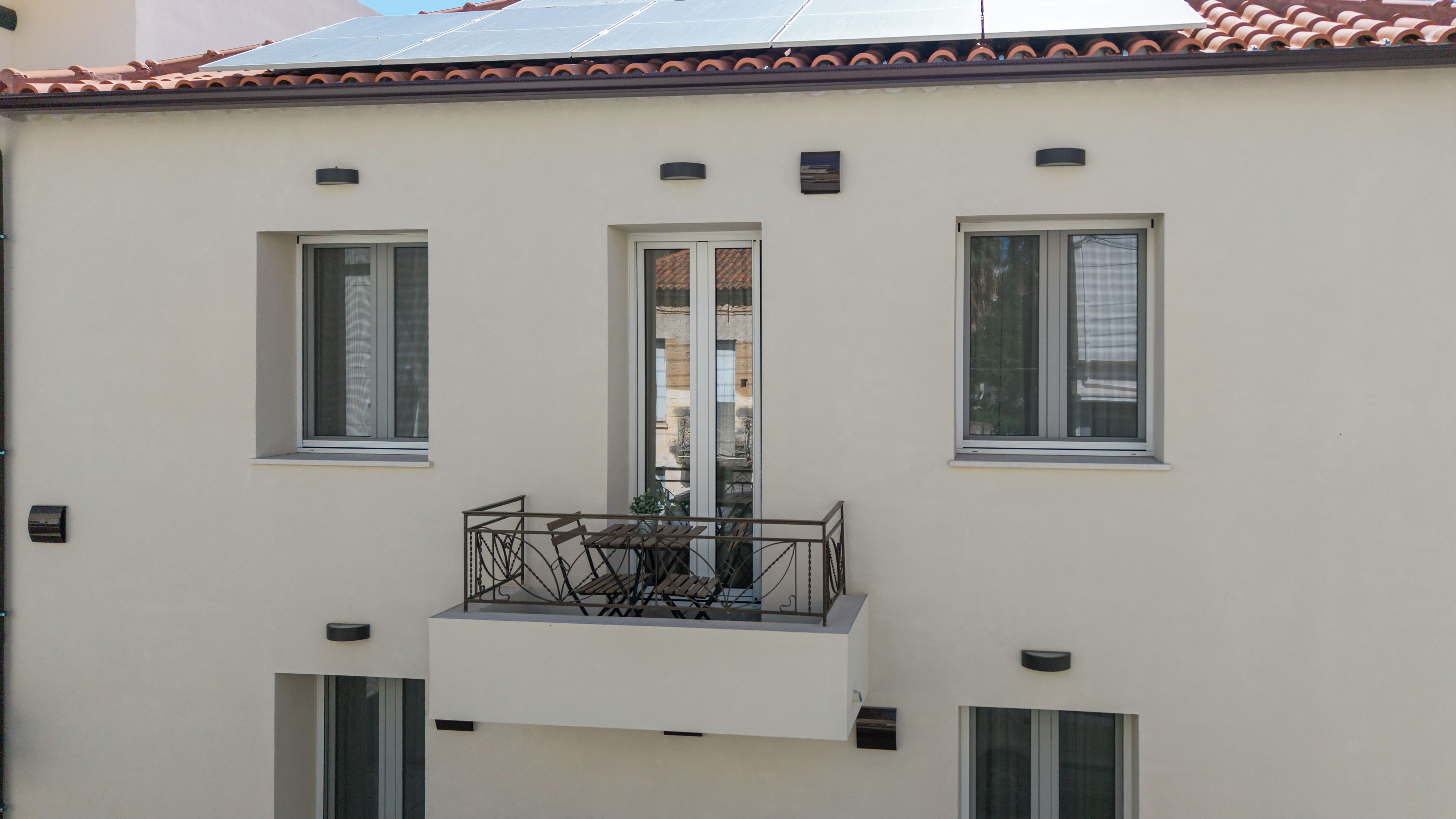 A building with three windows, a balcony with a table and chairs, and a solar panel on the roof.
