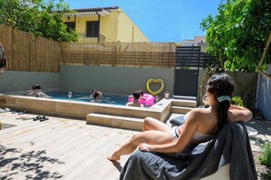 Zona de piscina con un deck de madera, cerca con valla de bambú y un edificio amarillo al fondo.