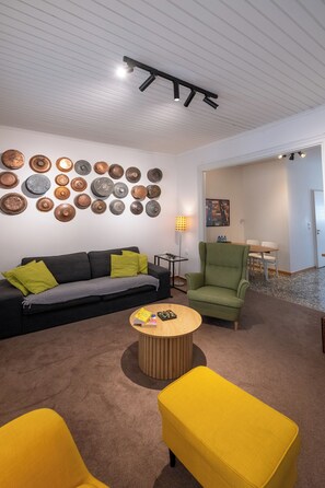 Una sala de estar moderna con un sofá negro, un sillón verde y un otoman amarillo. Una mesa de centro redonda de madera está en el centro.
