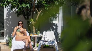 A person sitting on a white chair under a tree, with a table beside them containing a glass of orange juice, a glass of water, and a book.
