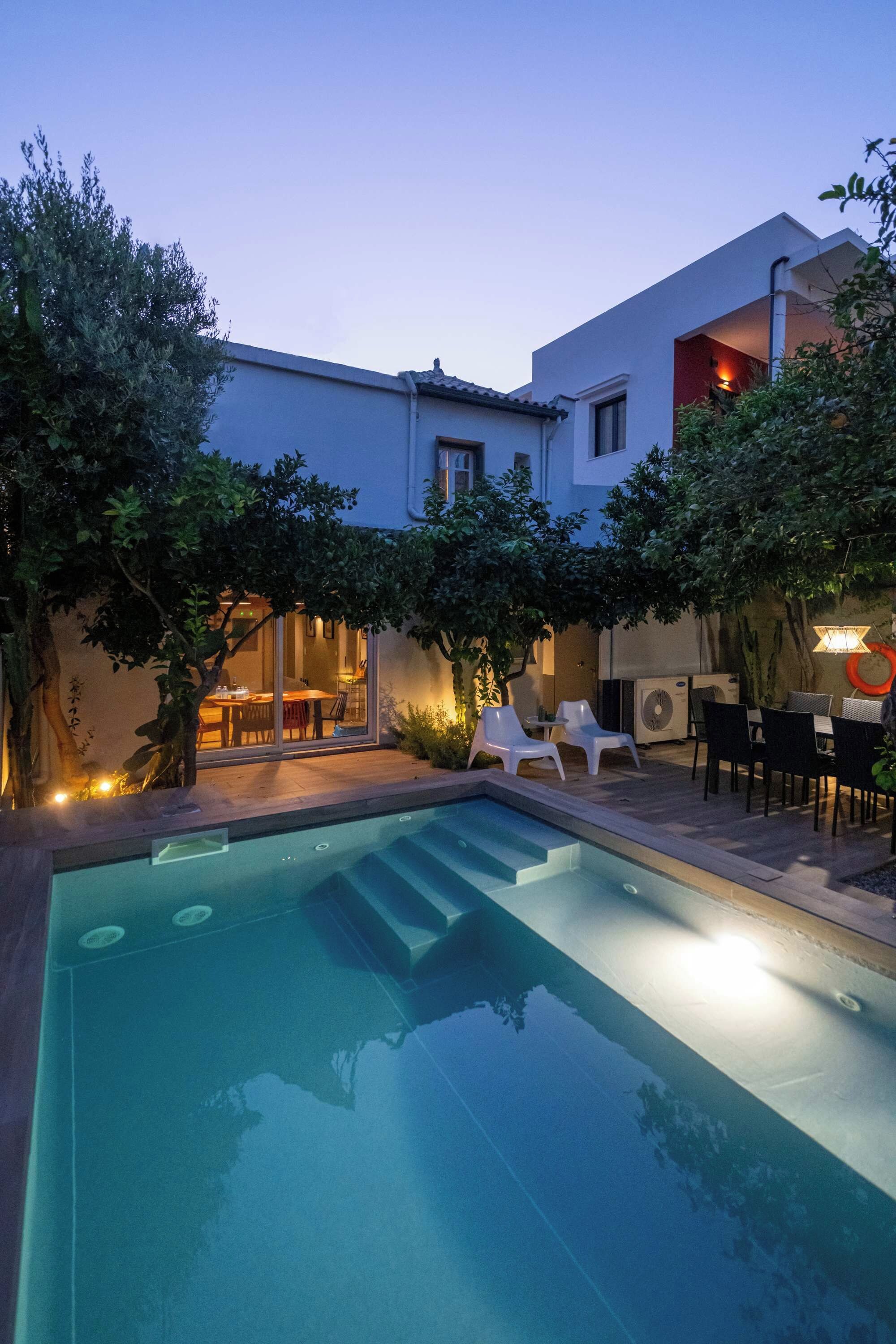 A swimming pool with lounge chairs and a table, surrounded by trees and a building with a tiled roof.