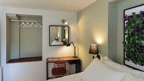 Una habitación de hotel con cama, escritorio y silla, espejo, lámpara y planta en la pared.