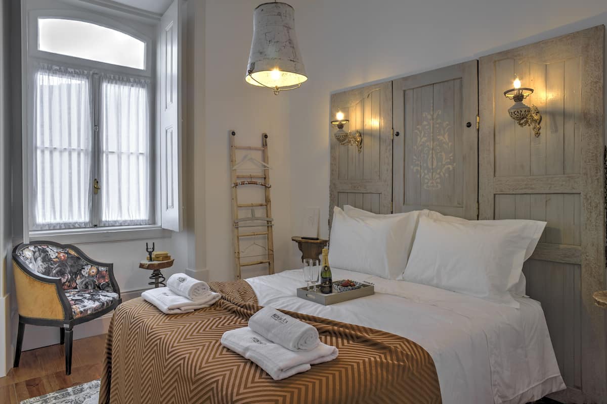 Uma cama bem arrumada com colcha estampada, uma mesa de cabeceira com garrafa e copos, uma cadeira com almofada florida e uma janela com cortinas.