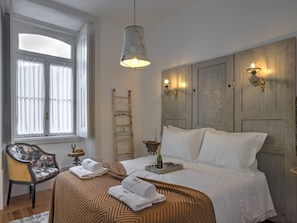 A neatly made bed with a patterned bedspread, a bedside table with a bottle and glasses, a chair with a floral cushion, and a window with curtains. - República Guest House (Coimbra)