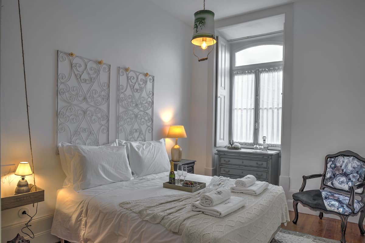 Cama bem arrumada com lençóis brancos, luminária de cabeceira e mesa de cabeceira com uma luminária. Janela com cortinas, cômoda e cadeira com almofada floral.