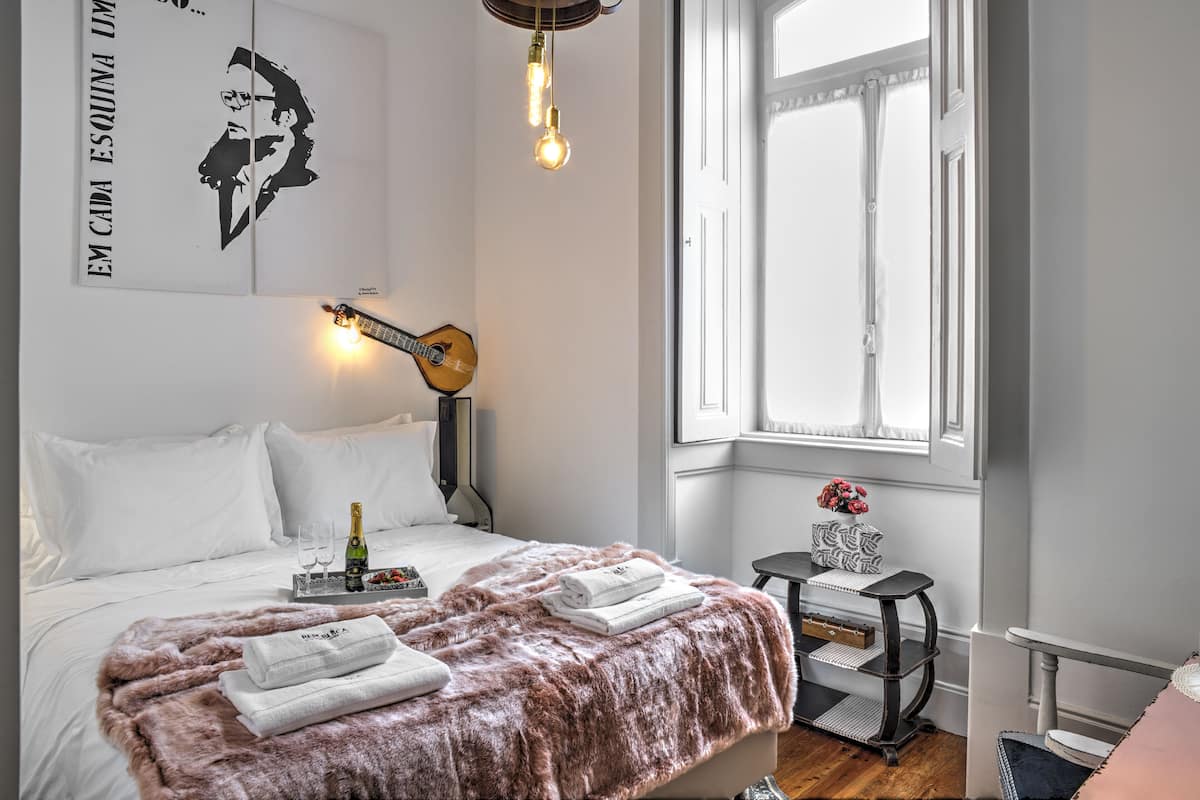 Quarto bem arrumado com uma cama, um violão, uma janela e uma mesinha com um vaso.