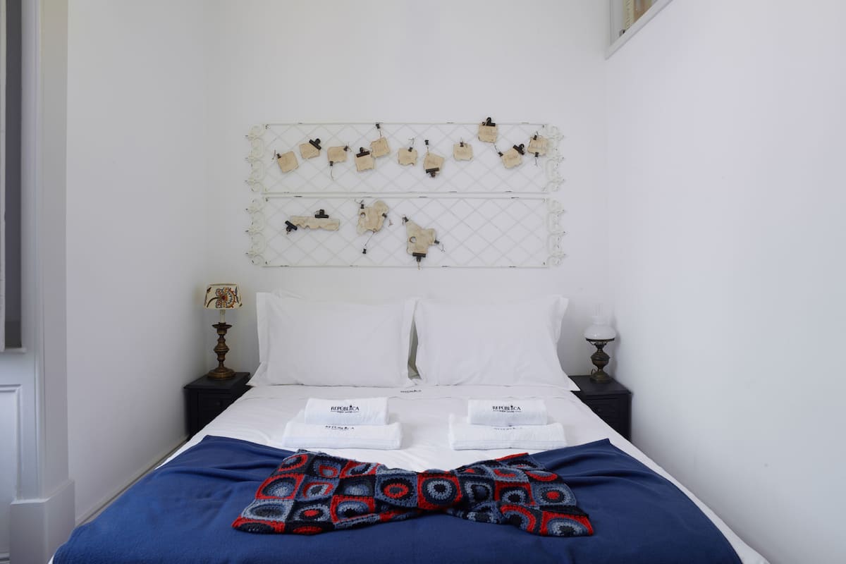 Uma cama bem arrumada com lençóis brancos e manta azul, uma prateleira fixada na parede com objetos decorativos, e uma mesa de cabeceira com um abajur e um pequeno objeto decorativo.
