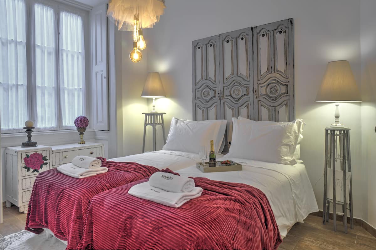 Quarto de hotel com cama, duas luminárias de cabeceira, um painel decorativo na parede, uma janela com cortinas leves e uma mesa de cabeceira com abajur e um vaso.
