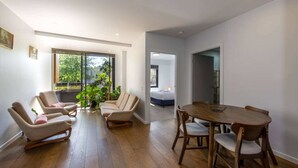 Ein modernes Wohnzimmer mit einem Holzesstisch, zwei beigefarbenen Sesseln und einem großen Fenster mit Blick auf Grünflächen.