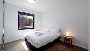 Ein Schlafzimmer mit einem großen Bett, einem Fenster mit Blick auf Grünflächen, einem Nachttisch mit Lampe und einer Deckenleuchte.