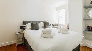 A modern bedroom with a large bed, bedside tables, a nightstand, a reading chair, and a bookshelf.