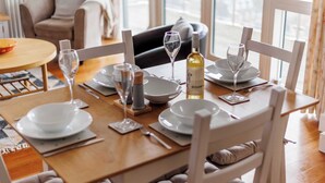 A dining table set with plates, glasses, cutlery, and a bottle of wine, with a sofa and a window in the background.