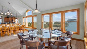 A spacious dining area with a glass table surrounded by leather chairs, large windows offering a view of the landscape, and a kitchen area with wooden cabinets and a hanging light fixture. - Premier Soundfront Dog Friendly Oasis w/ Pool, Hot Tub, Elevator, and Much More! (Avon)